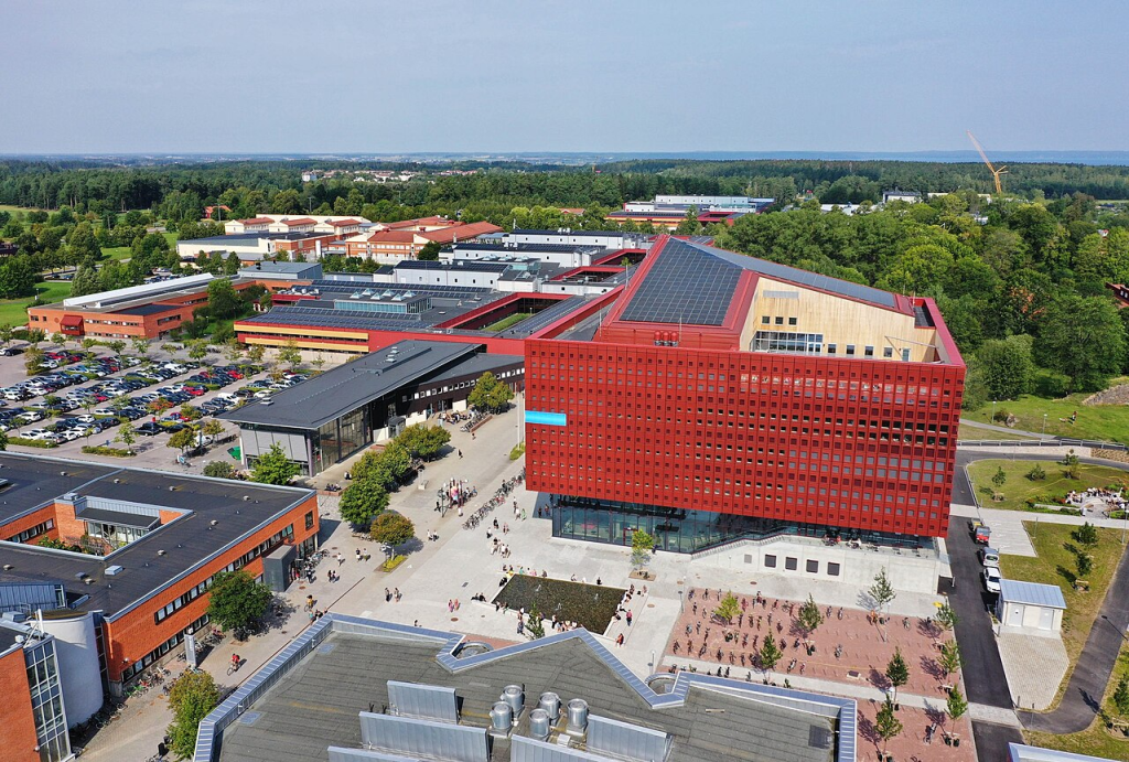 A picture of the campus of Linkoeping University, taken from above. We see several academic buildings set against a forested background. In the far distance, we can see the larger town stretching off towards the horizon. The primary academic building is a large modern red building, with solar panels on the roof and a rooftop courtyard.campus valla linköping aboveErik And30 - Own workCC-By-SA 4.0